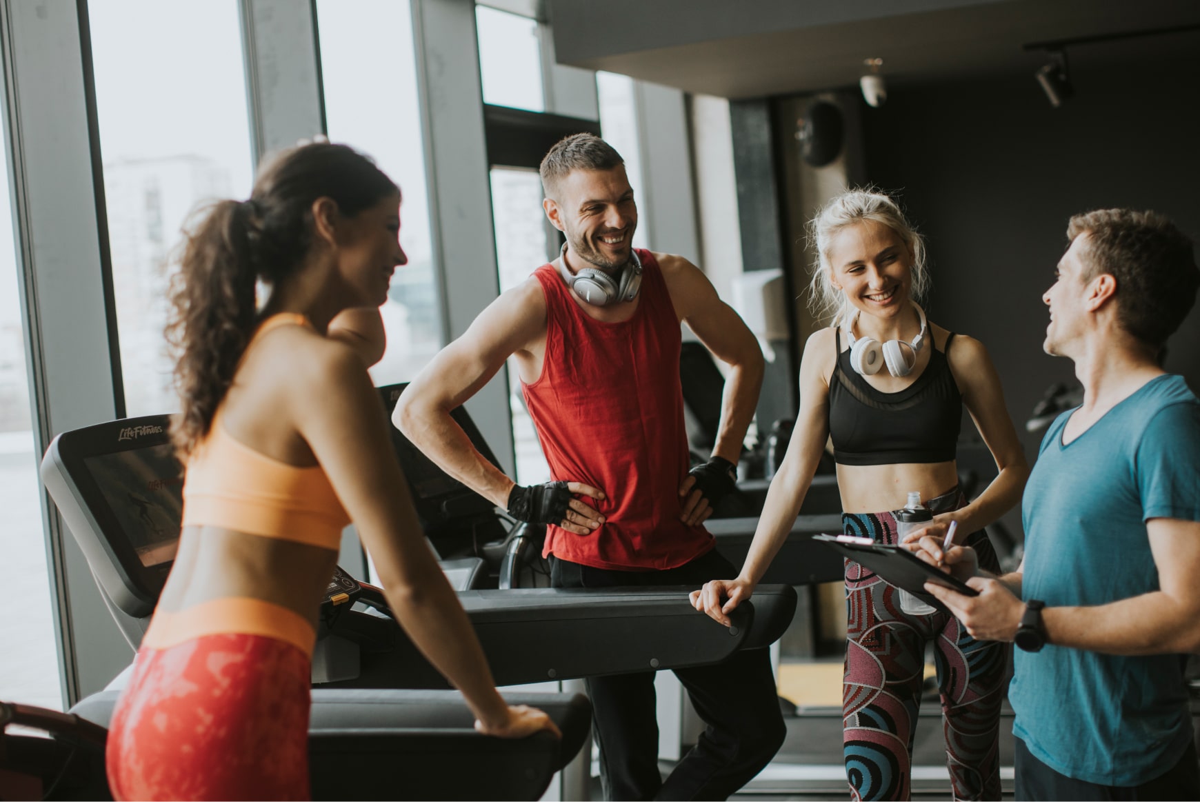 Trainer with a clipboard chatting with 3 smiling gym-goers on treadmills after a workout.