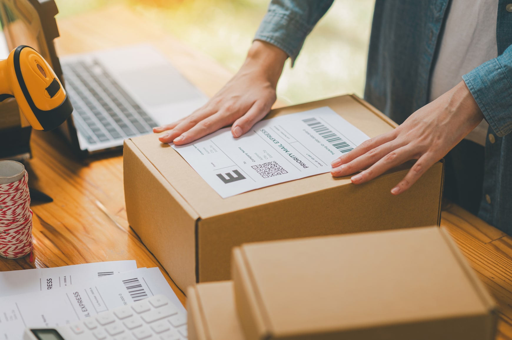 A small business owner in an apron carrying packages while managing online orders on a laptop.